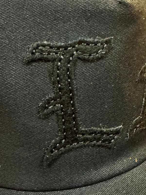 Close-up of a black embroidered letter 'L' on a black fabric cap, showing the texture of the thread and woven material.