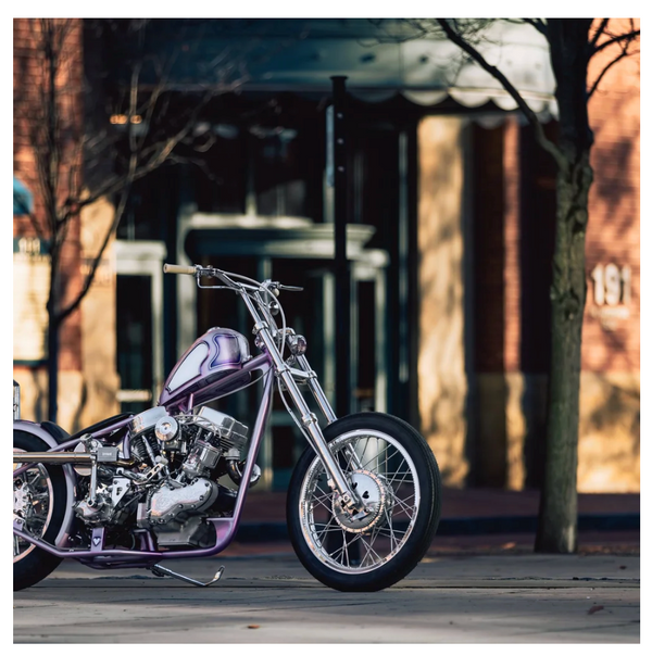 A custom purple chopper motorcycle with a chrome engine and front fork, parked on a city street in front of a blurred brick building.