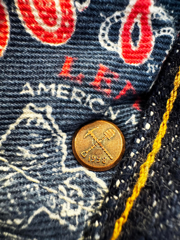 Close-up of a vintage denim garment showing a bronze-toned metal button engraved with an anchor and the year 1998, alongside red and white embroidered graphics and yellow contrast stitching.