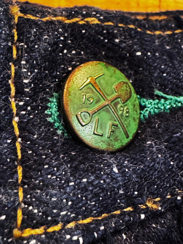 A close-up of a round, vintage green metal button with '1948 DLF' and crossed tools engraved, pinned onto a black denim fabric with yellow and green stitching.