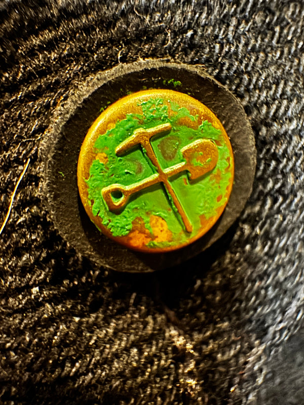 A close-up of a round vintage button with a yellow-gold background and a green patina, featuring a raised crossed pickaxe and shovel symbol. The button is set in a dark circular frame and mounted on a dark, textured fabric.