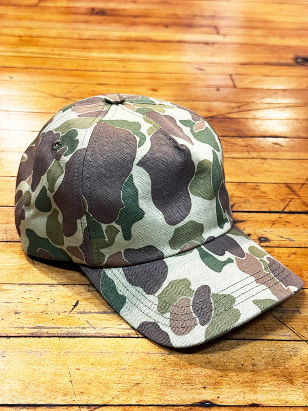 A baseball cap with a classic camouflage pattern in shades of green, brown, and beige, resting on a wooden plank floor.