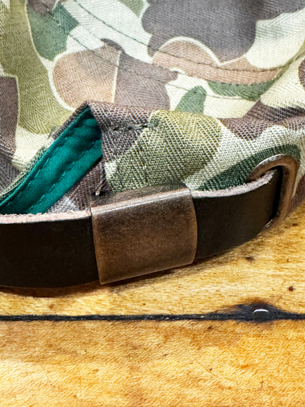 Close-up of a camouflage-patterned fabric item with a dark brown leather strap and a rectangular metal buckle, resting on a textured tan surface.