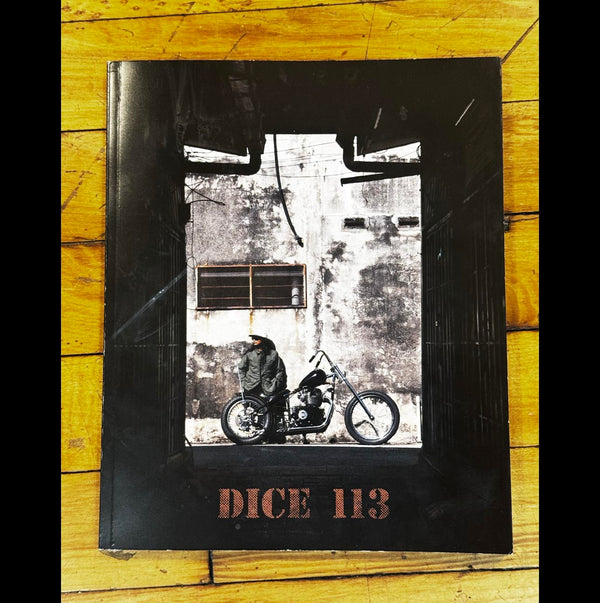 A square black book or album cover titled 'DICE 113' lies on a yellow wooden surface. The cover features a monochrome photograph of a person sitting on a motorcycle in front of a weathered wall, framed by a thick black border.