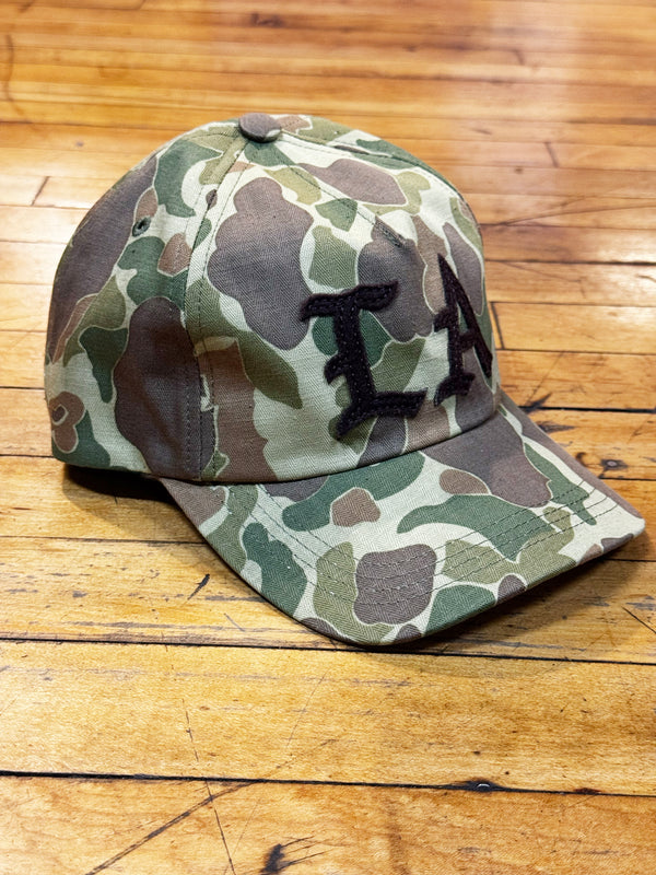 A baseball cap with a green, brown, and beige camouflage pattern, featuring a black embroidered 'LA' on the front, resting on a wooden floor.
