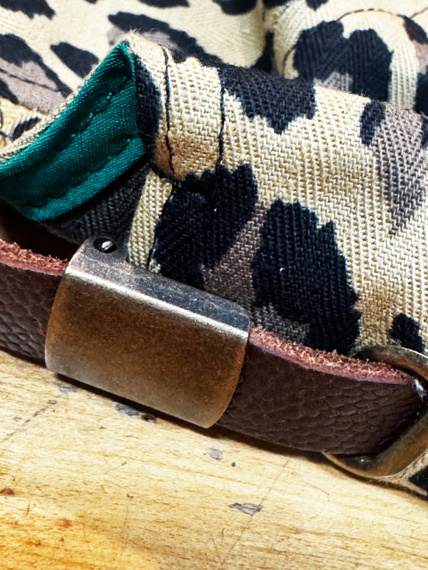 Close-up of a dog collar featuring a brown textured leather strap, a rectangular bronze-toned metal buckle, and a woven fabric strap in a beige, black, and brown camouflage pattern with a teal lining.