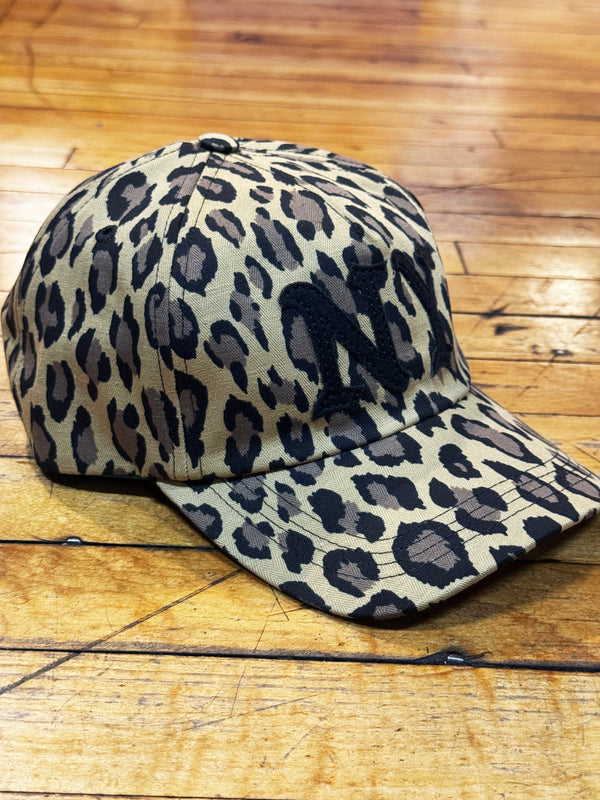 A baseball cap with a leopard print pattern in beige, brown, and black, resting on a wooden floor.